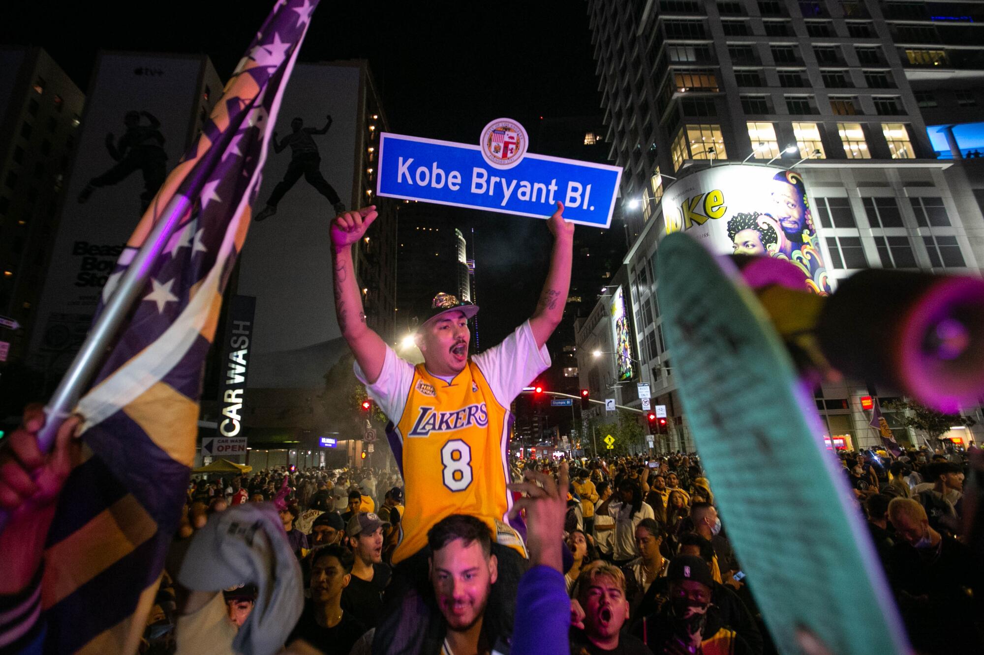 A man in a gold Lakers 8 jersey on the shoulders of another man holds a Kobe Bryant Boulevard street sign