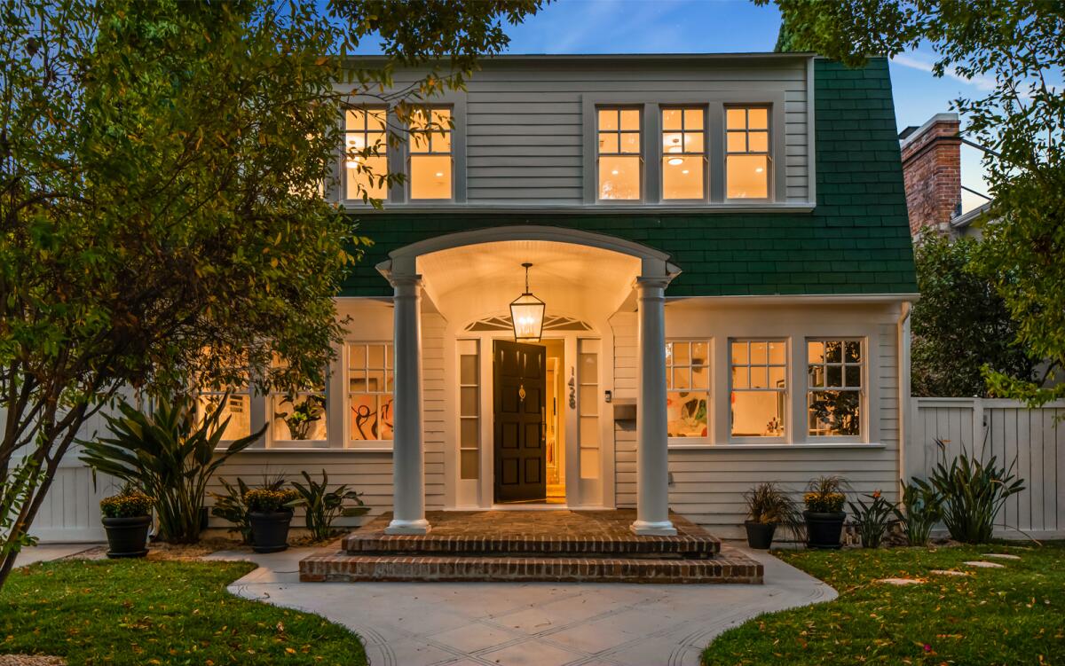 A two-story white house with a pair of columns at the entrance is shown in the evening with its interior lights on.