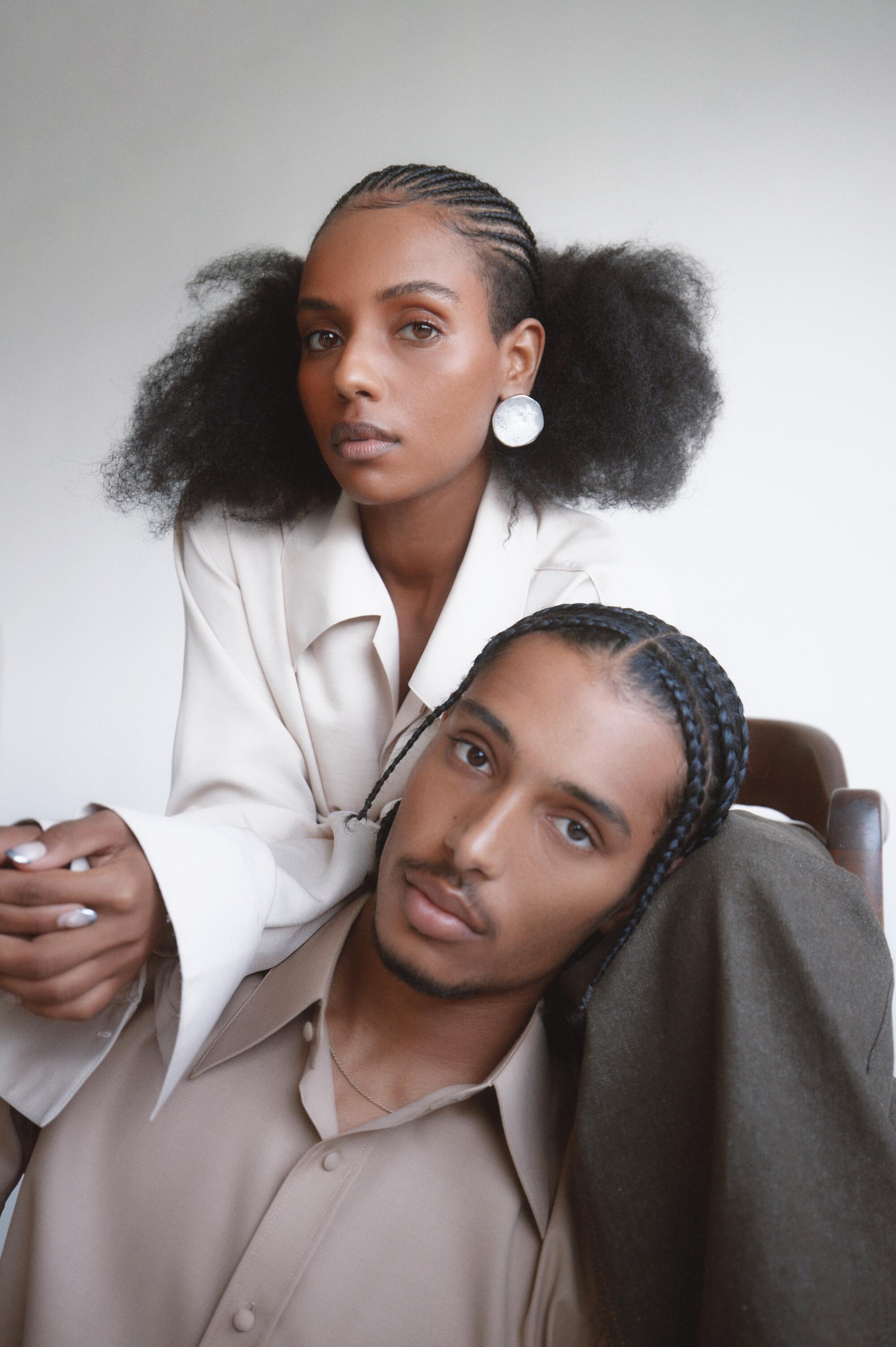 A male model wearing a beige collared shirt leans his head against the leg of a female model wearing a white collared shirt.