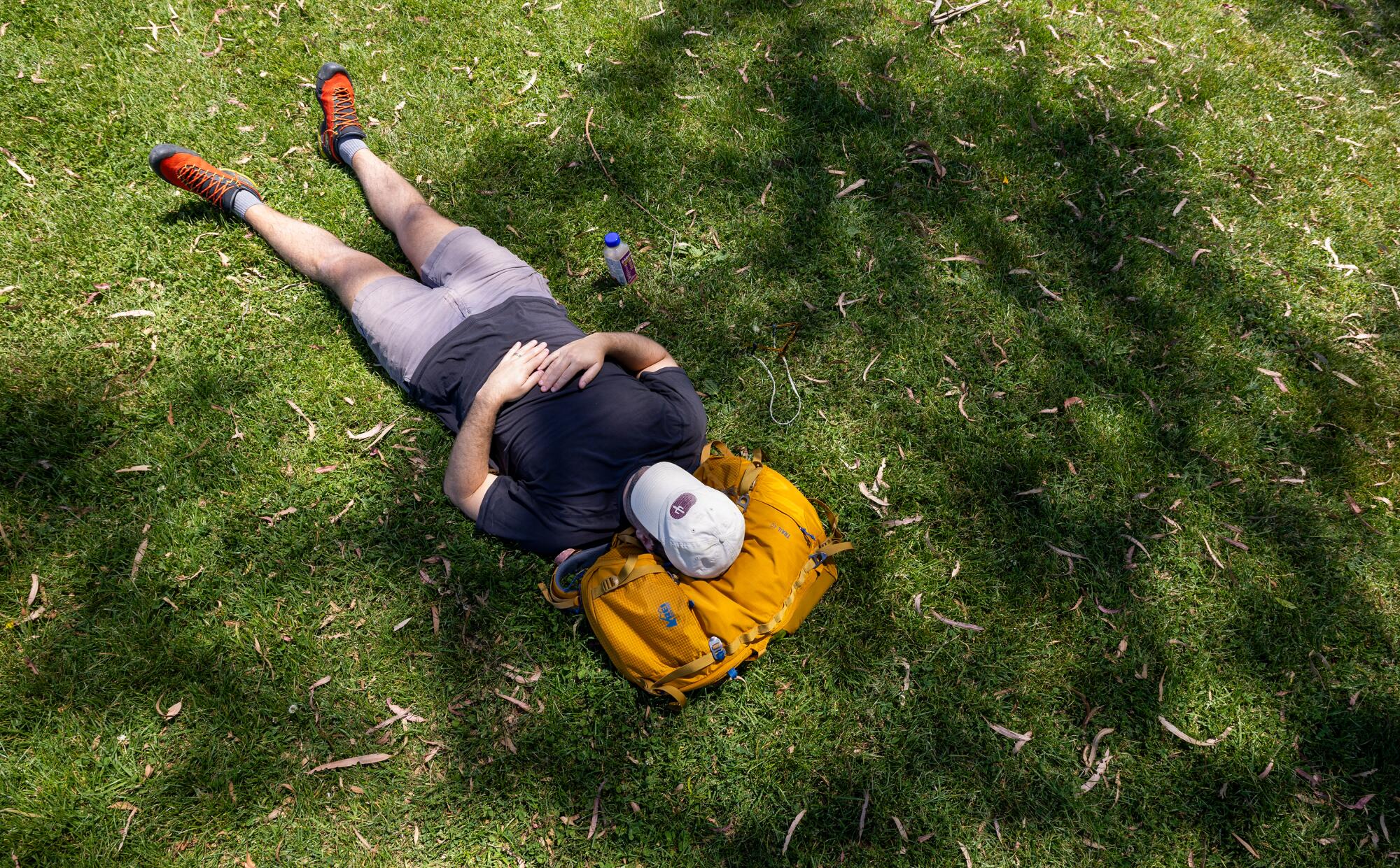 Un hombre está tumbado en el césped, usando su mochila amarilla como almohada.