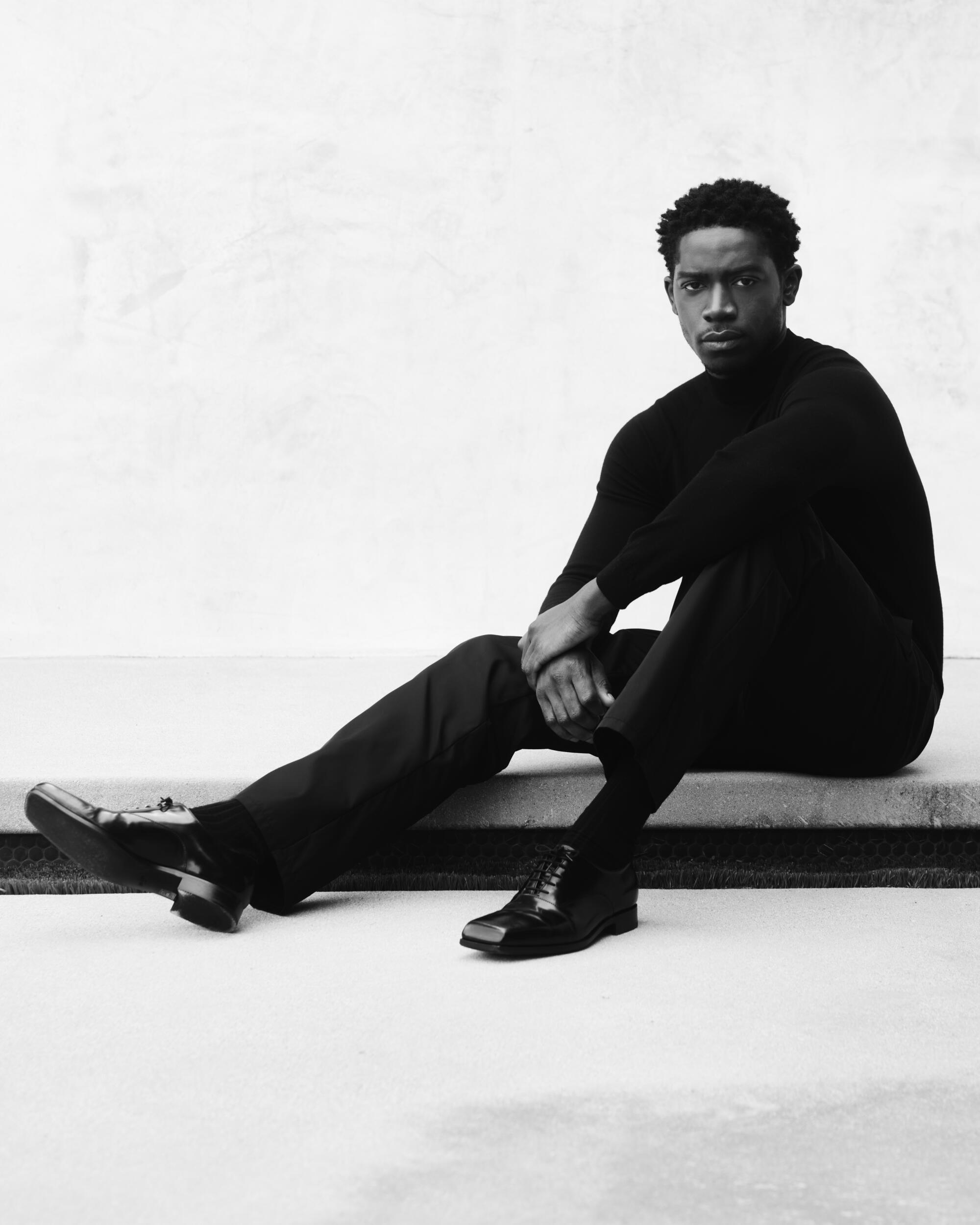 In a black-and-white photo, Damson Idris, dressed in black, sits on the ground with one leg outstretched. 