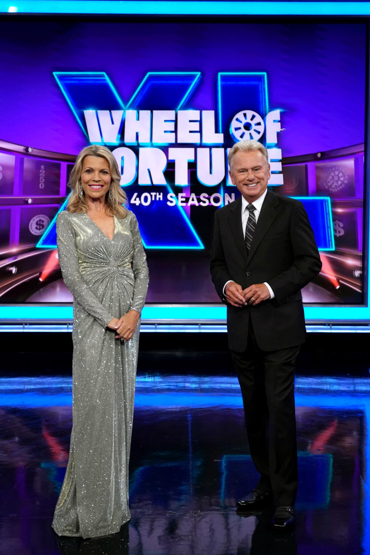 Vanna White, in a shimmery silver gown, and Pat Sajak, in a suit, stand on the "Wheel of Fortune" stage.