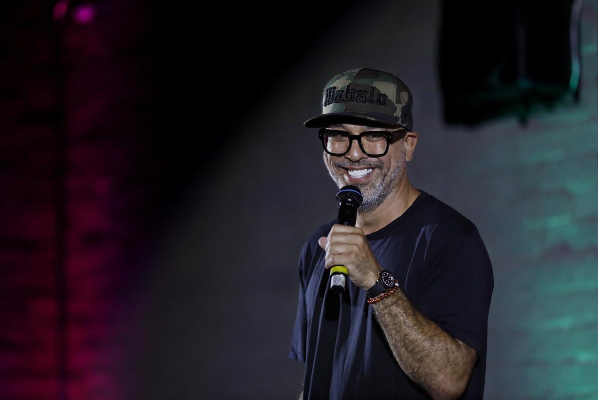 A man in a black T-shirt and baseball hat holds a microphone to his mouth.