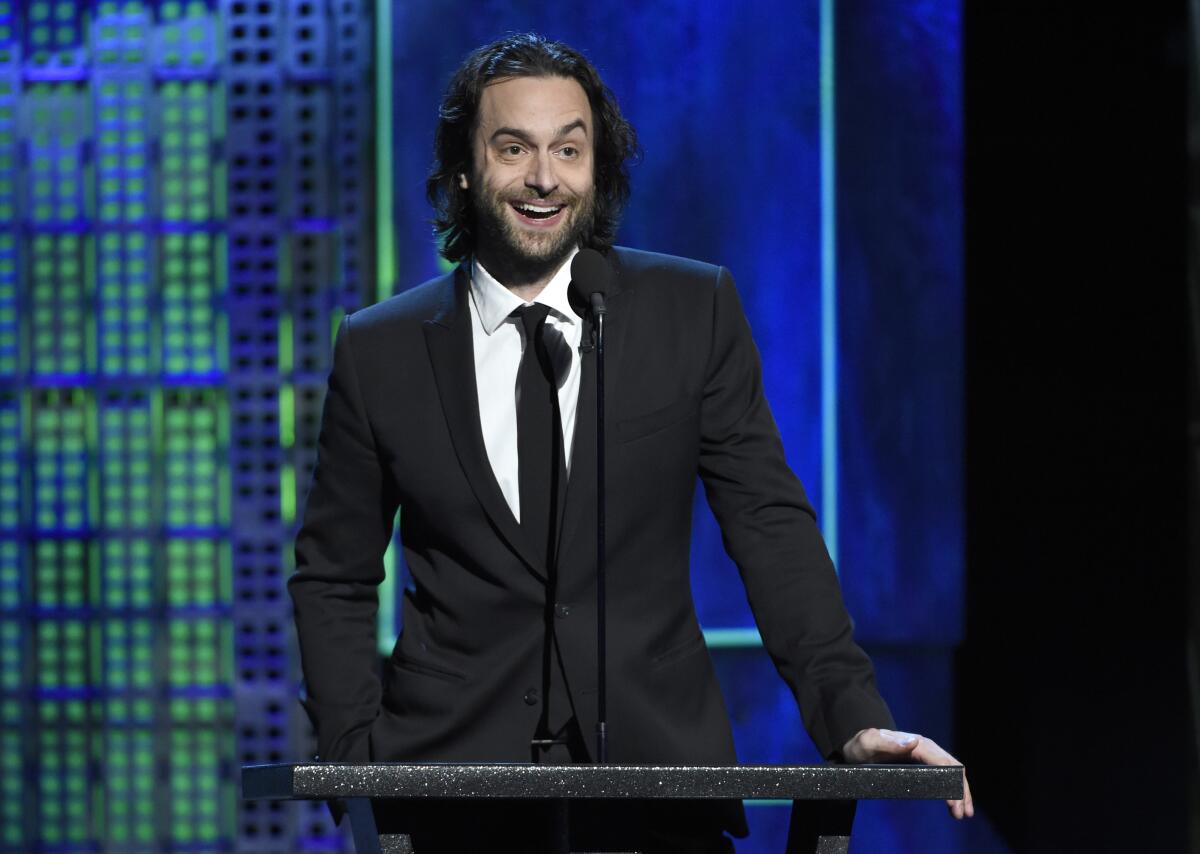 Chris D'Elia stands at a microphone wearing a dark suit