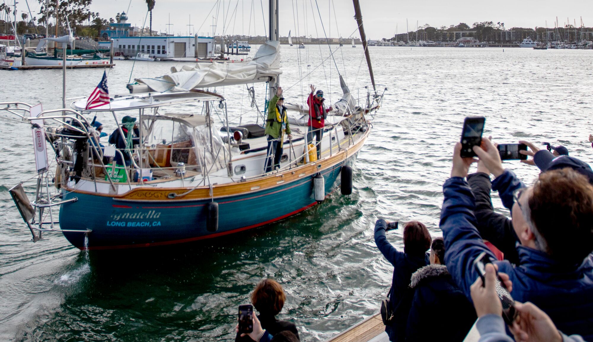 A ship sets sail in a harbor while a group of people wave and record on their cell phones. 
