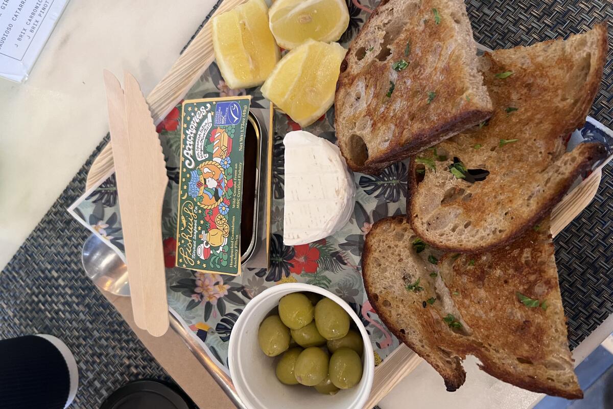 A tin of fish on a platter with herbed toast, a half-moon of brie, lemon wedges and green olives. 