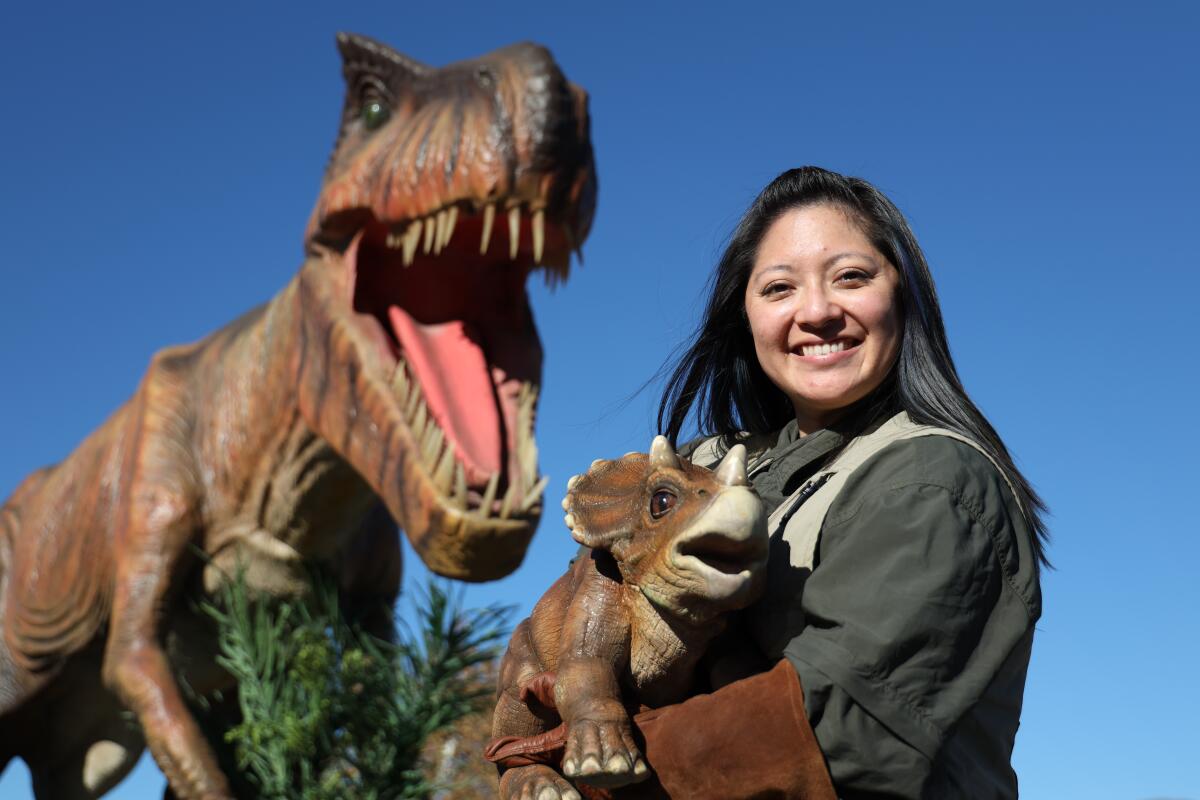 A dinosaur trainer holds a baby triceratops, posing in front of a T-Rex, at a Jurassic Quest event.