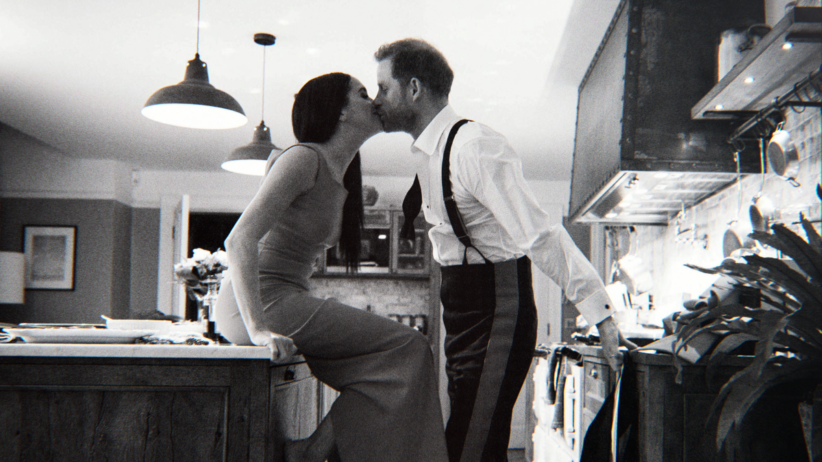 A woman sitting on a kitchen island and a man in suspenders kiss.