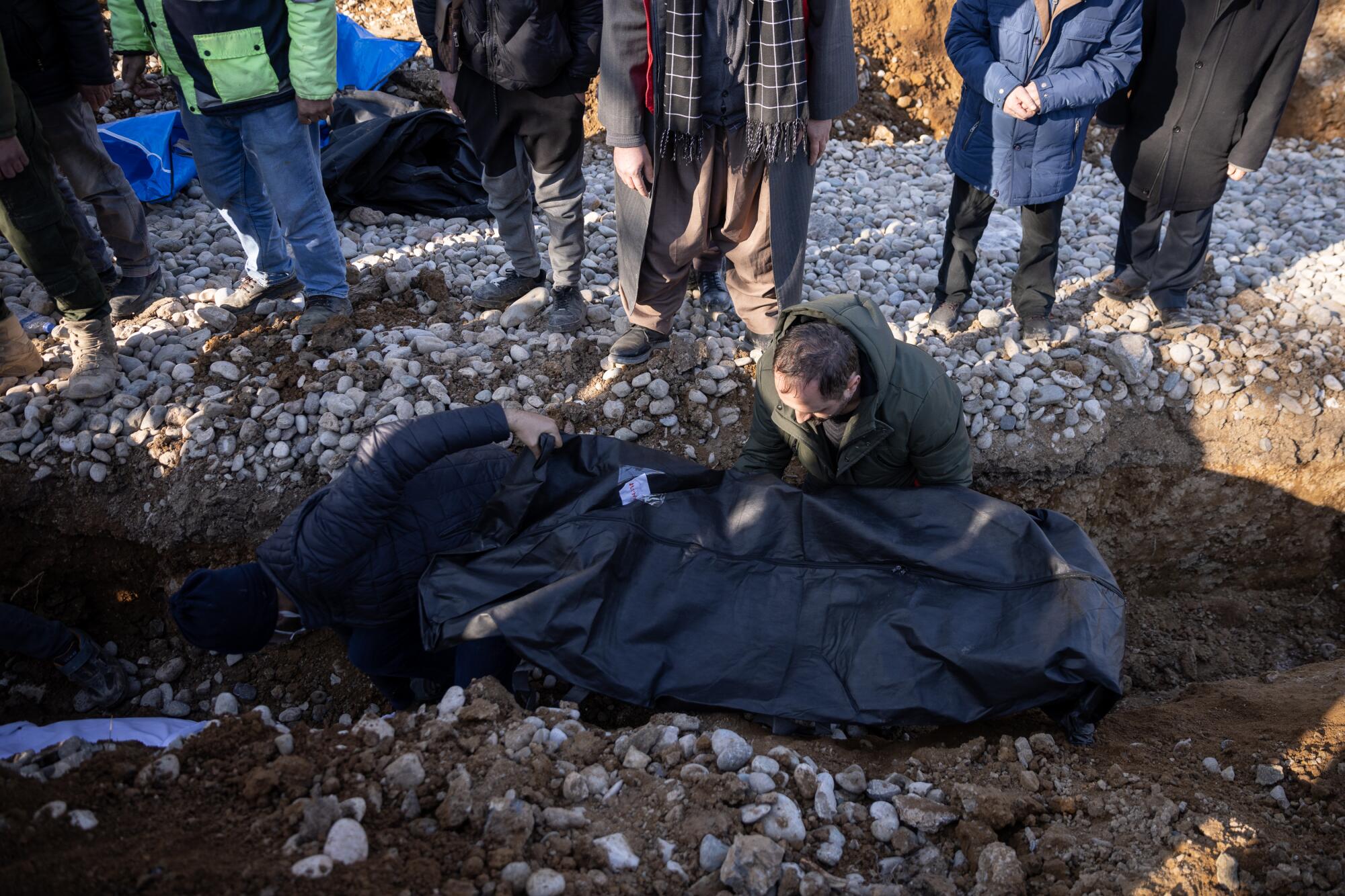 Men placing a body in a black body bag in a dirt trench as mourners stand at the edge 