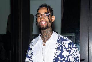 PnB Rock smiles while wearing glasses, a chain and a blue and white Hawaiian shirt 