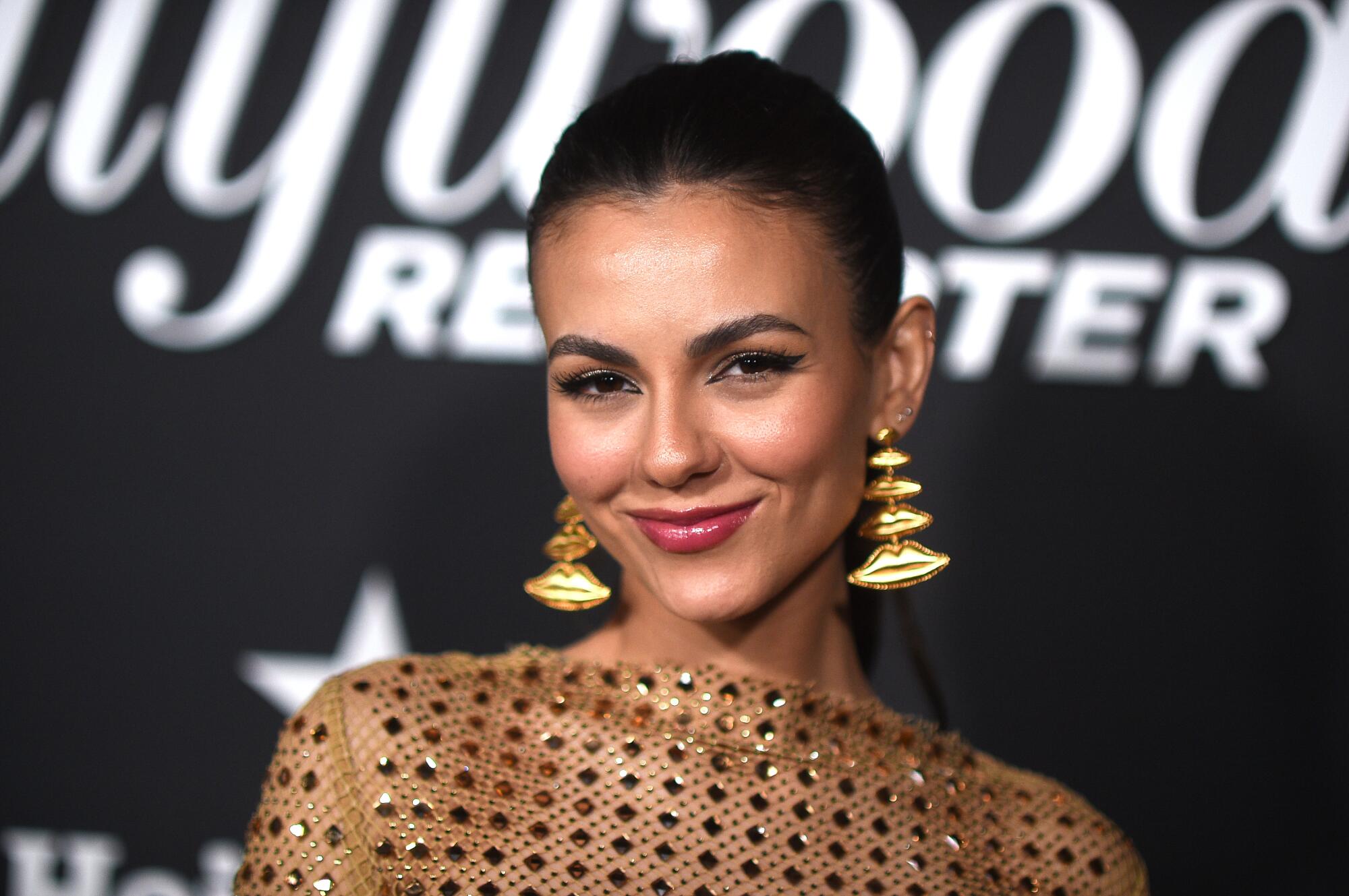 Victoria Justice, shown from the shoulders up, wears a sequined gold dress and statement earrings. 