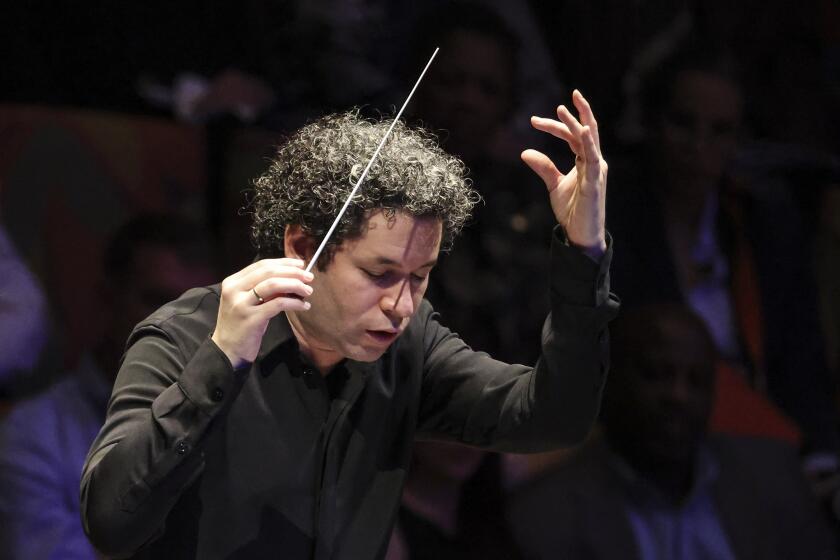 Gustavo Dudamel, wearing black and looking serioius, holds up a white baton as he conductts