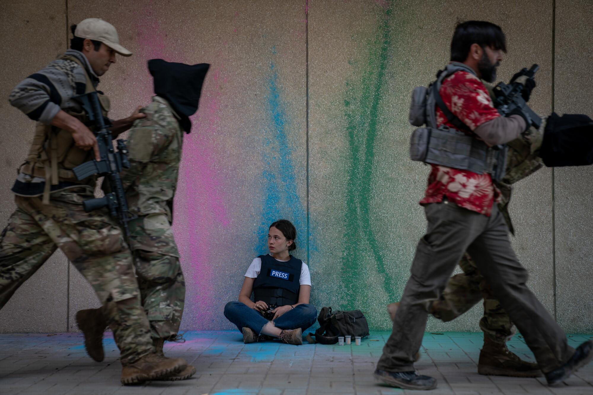 A press photographer sits on the ground as soldiers march by with a detainee.