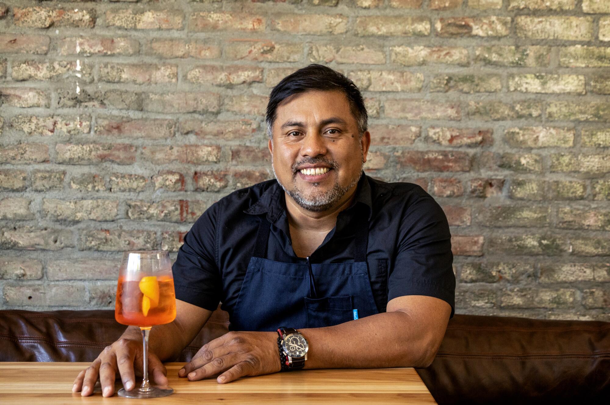 Man wearing an apron seated in front of exposed brick with hand on a wine glass 