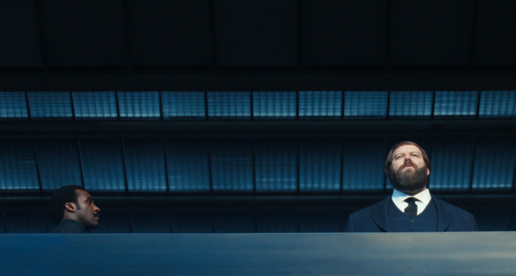 A man seen from the side looking at a bearded man standing and looking over a walkway.