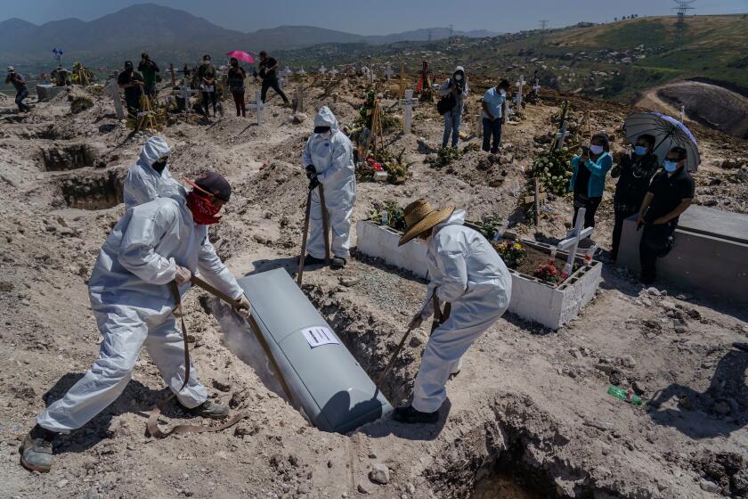 TIJUANA, BAJA CALIFORNIA -- MONDAY, APRIL 27, 2020: Cemetery workers lower the casket of Juan Velasco, who died of COVID-19 symptoms, as his family, to the right, watches the burial at the Municipal Pantheon 13 cemetery, in Tijuana, Mexico, on April 27, 2020. (Marcus Yam / Los Angeles Times)