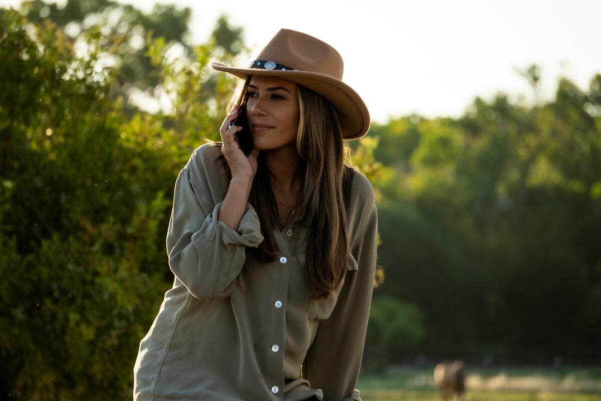 A woman in a brown cowboy shirt and brown hat sits in a wooded area and holds a cellphone to her ear.