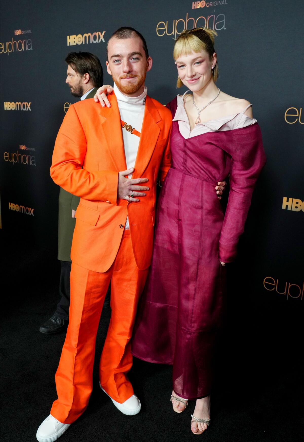 Angus Cloud poses in an orange suit next to Hunter Schafer, who has her arm thrown over his shoulder