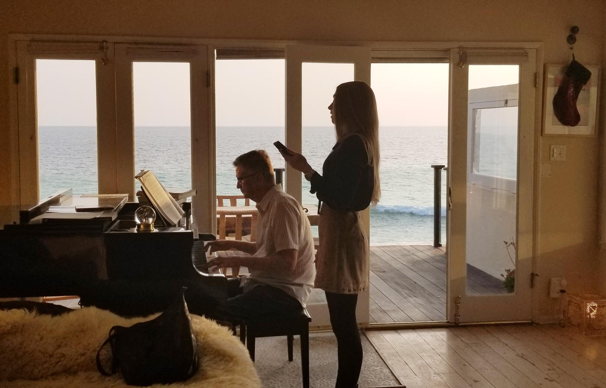 Mark Sawusch plays the piano at his Malibu beach house, with Anna Moore behind him.