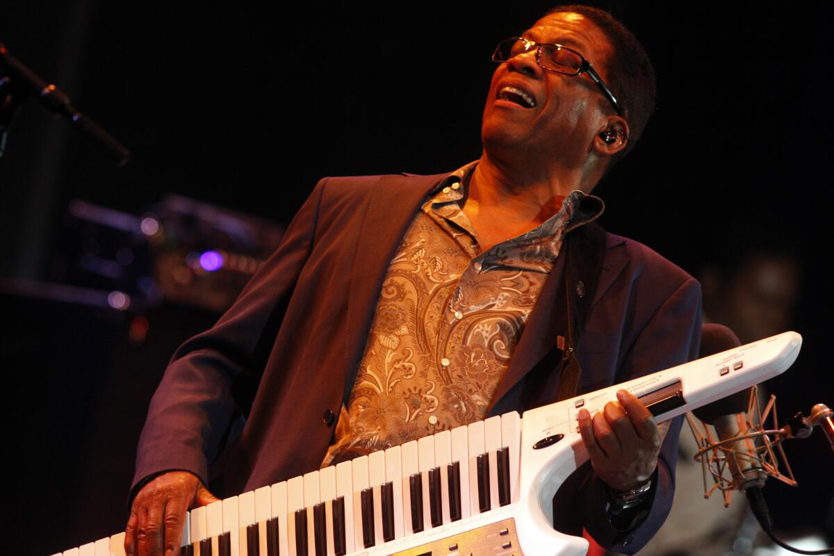 Herbie Hancock, wearing a paisley shirt and a black blazer, gets down on stage while playing a keytar.