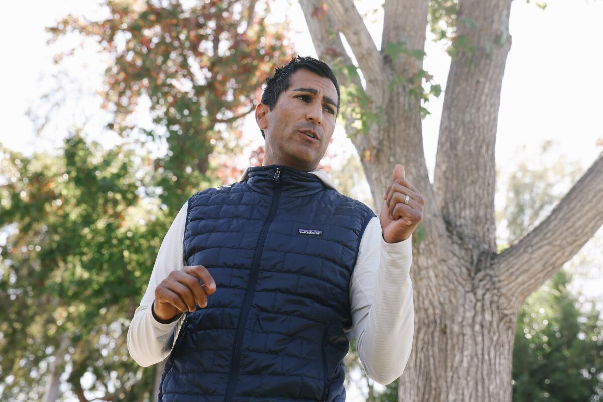 A man speaks outdoors with trees in the background.