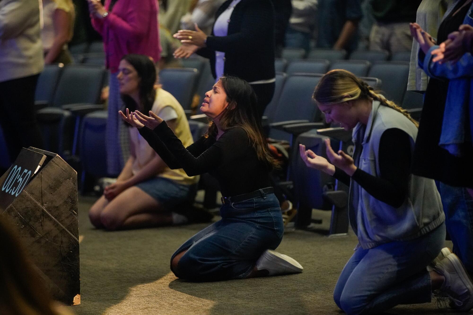 Several worshipers kneel in prayer, their hands outstretched, on a carpeted chapel floor.