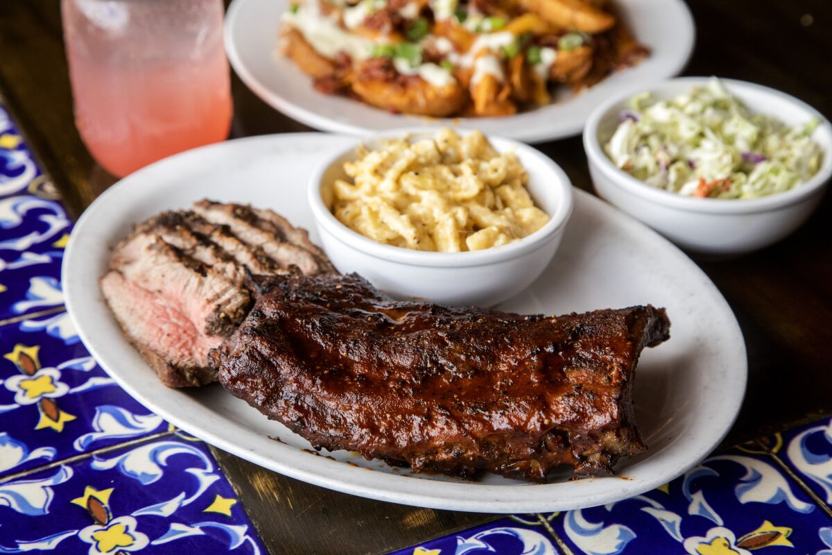 Plates of ribs, tri-tip, mac and cheese, coleslaw and cheese fries next to a pink drink in a Mason jar.