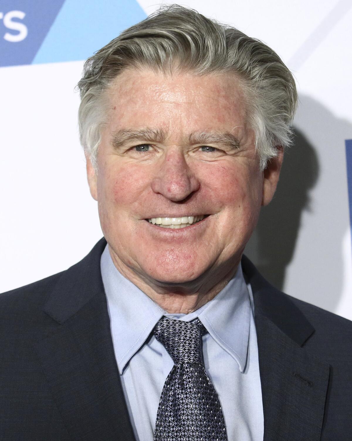Treat Williams with gray combed hair smiles while wearing a black suit with a light blue shirt and gray tie. 