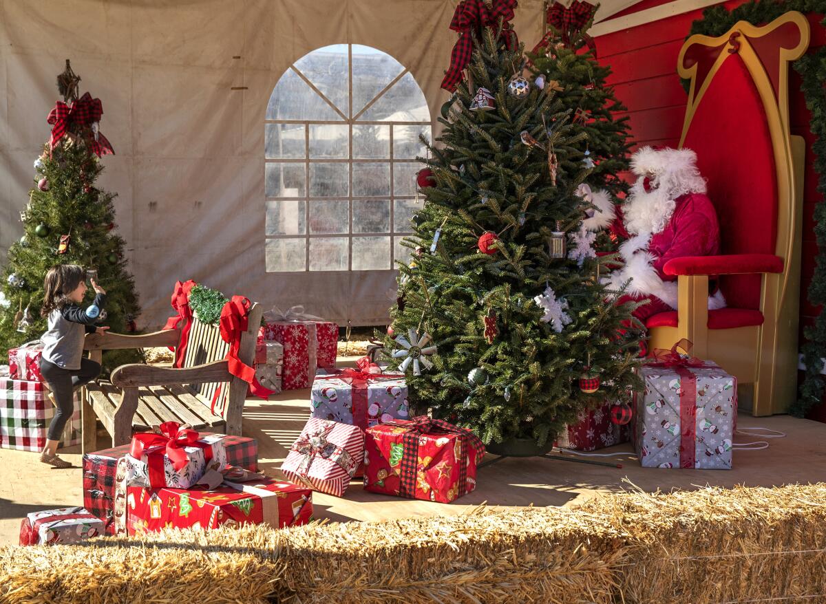 Zoie Dabit, 4, waves as she climbs onto a bench. She's separated from Santa by piles of gifts and a Christmas tree.