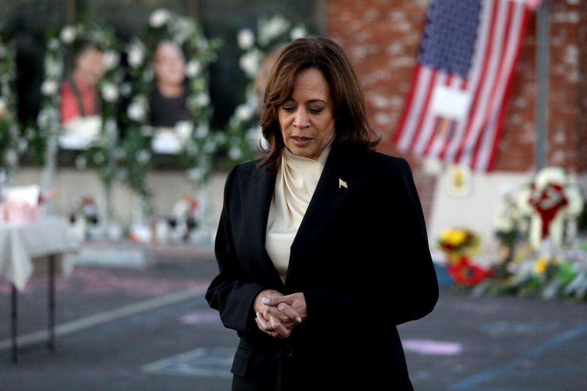 Kamala Harris bows her head and clasps her hands while speaking near flower memorials