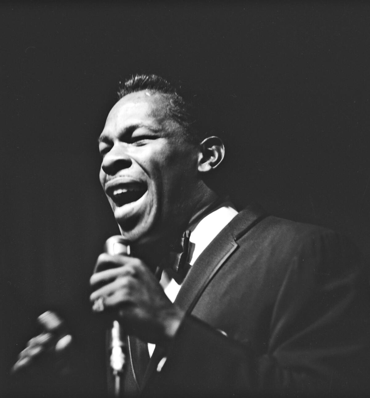 Wearing a suit and tie, Lloyd Price sings into a microphone.
