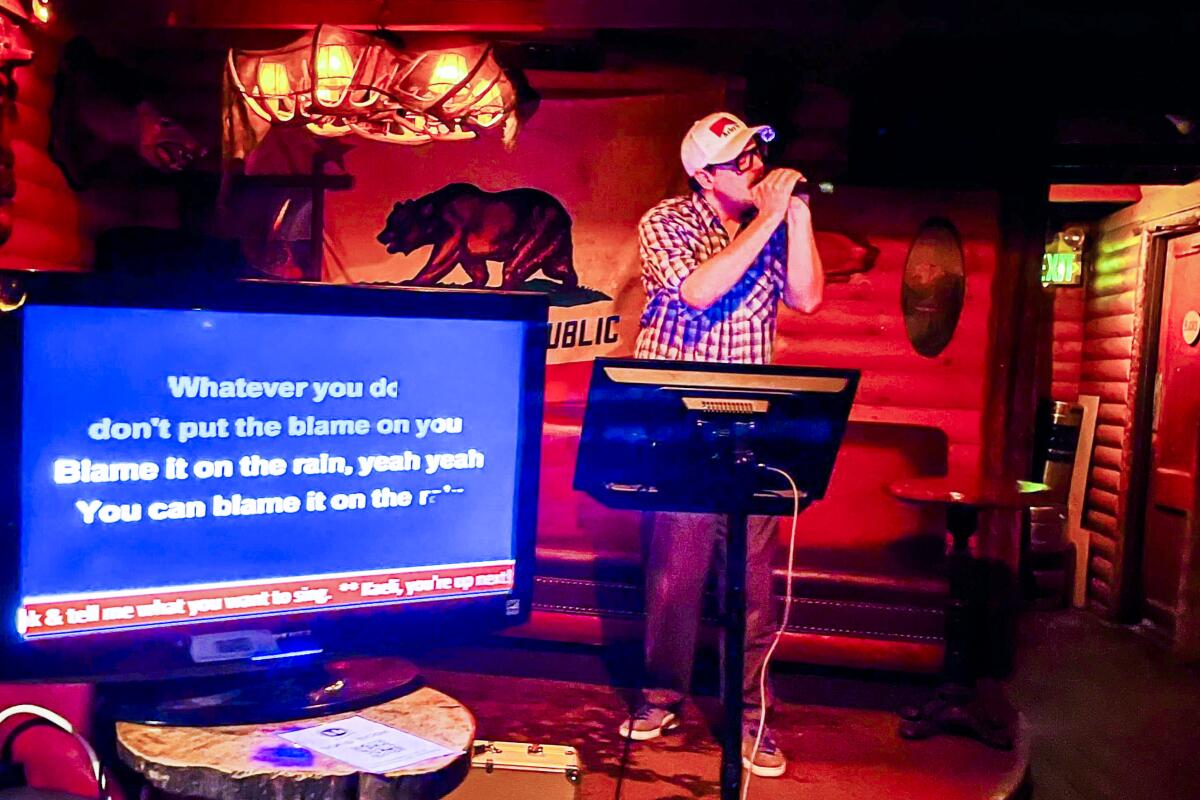 A man in a ball cap sings into a microphone next to a screen with the lyrics to a song.