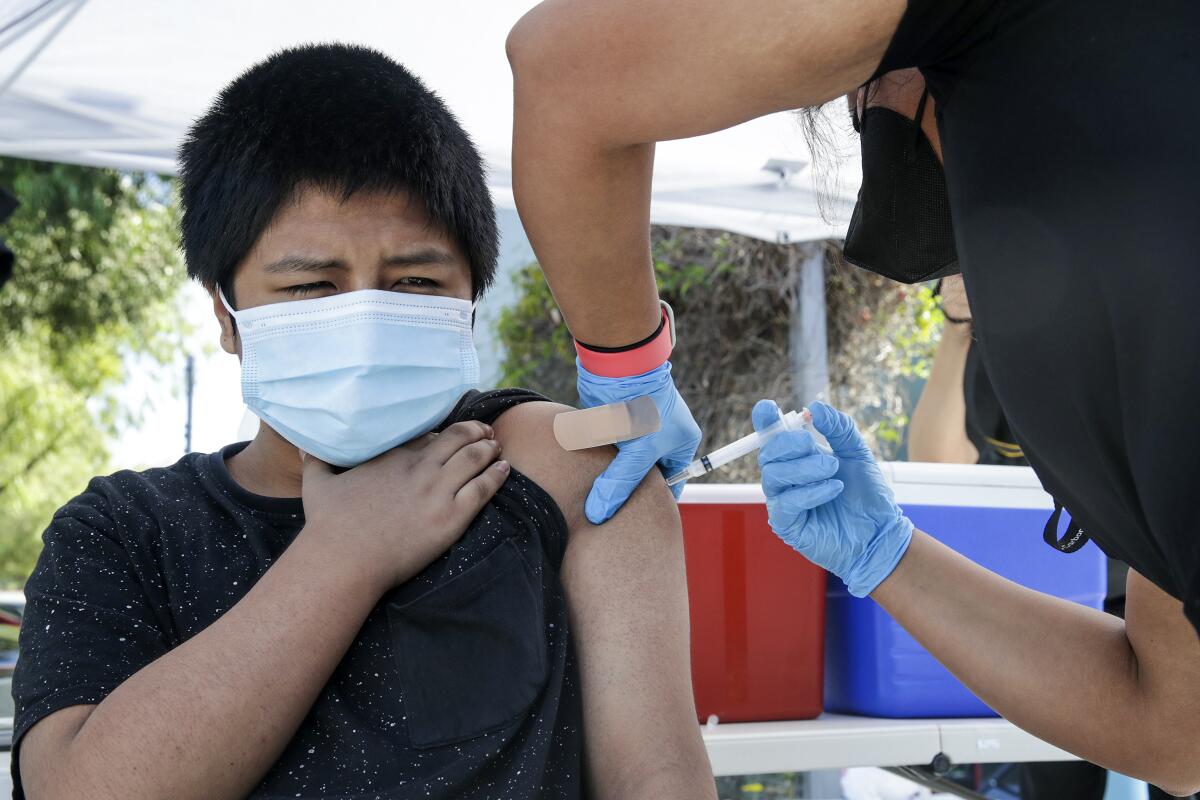 A masked boy holds a hand up to his neck as he gets a shot in the arm 