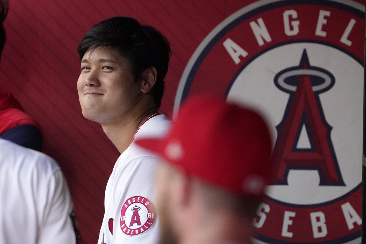 Shohei Ohtani stands and smiles in the dugout as he waits to hit. 