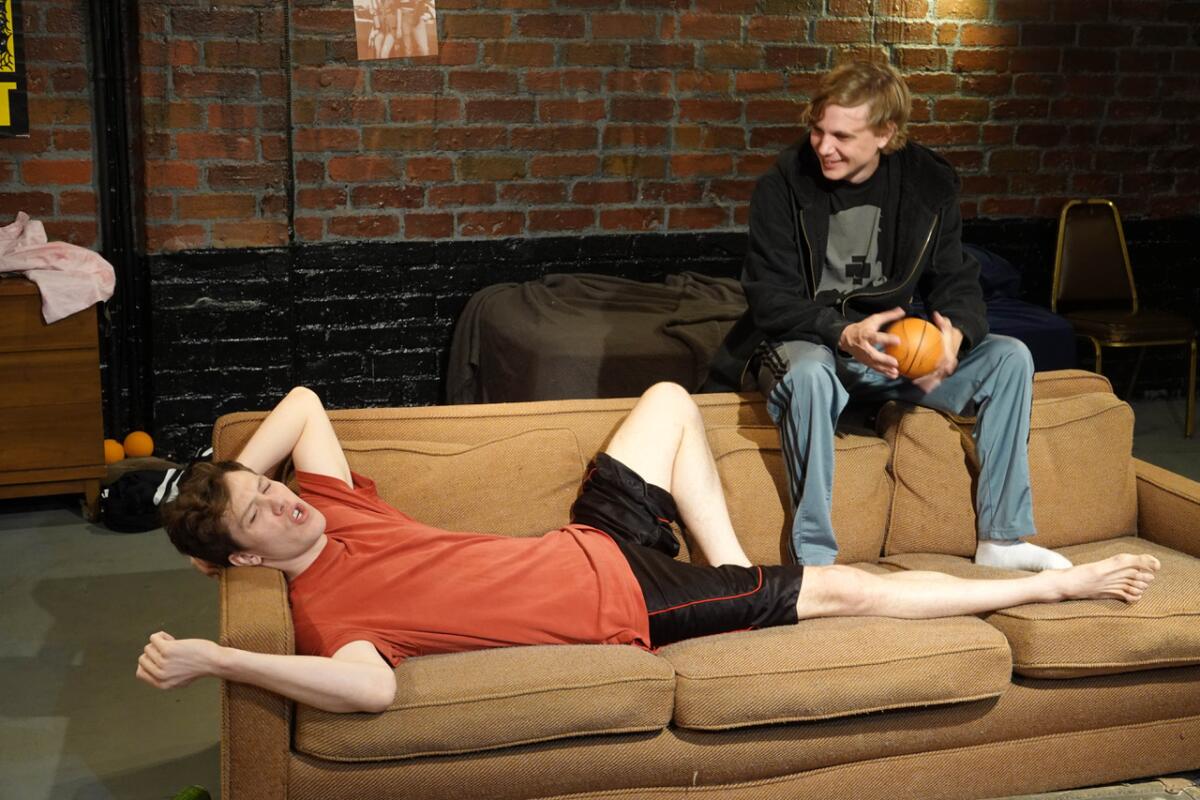 One man lies on a couch and another sits on the back of it, holding a small basketball.