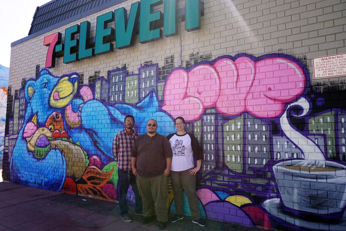 From left: Co-founder of Love City Heights Carlos Quezada, artist GMONIK and Danielle Iwatsu stand in front of the "Bear Essentials" mural on the wall of a 7-Eleven in City Heights on Oct 1, 2019.