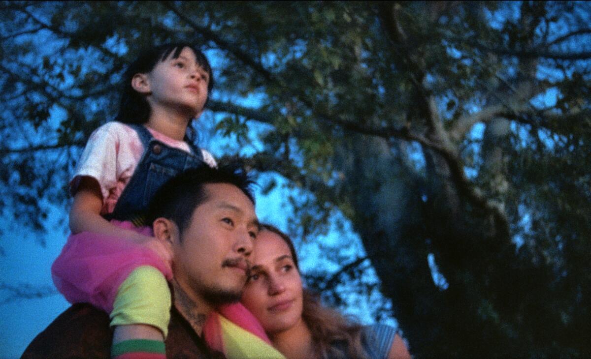 A man, with a child on his shoulders, and a woman stand in front of a tree in the movie "Blue Bayou."