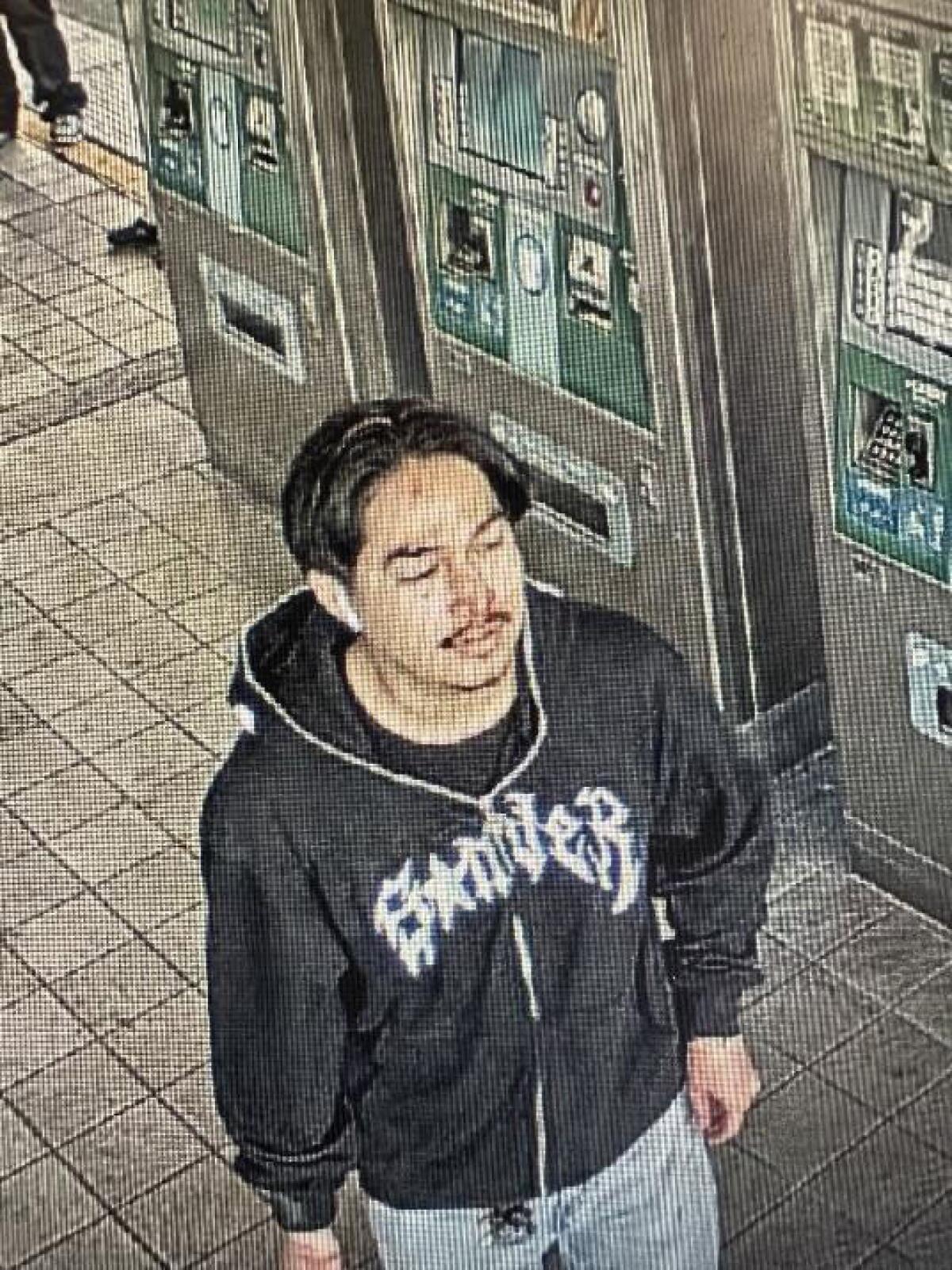 A man with dark hair and a black sweatshirt with white lettering walks past ticket machines on a train platform.