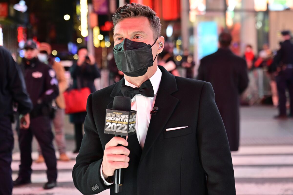 Ryan Seacrest, wearing a black tuxedo, tie and mask, holds a microphone.