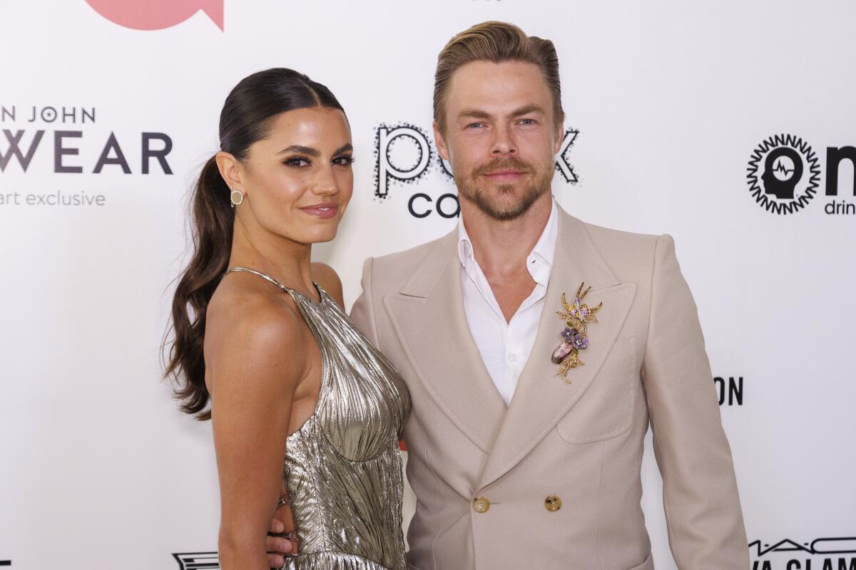 A woman in a silver dress stands with a man who wears a beige blazer with a gold broach and white shirt