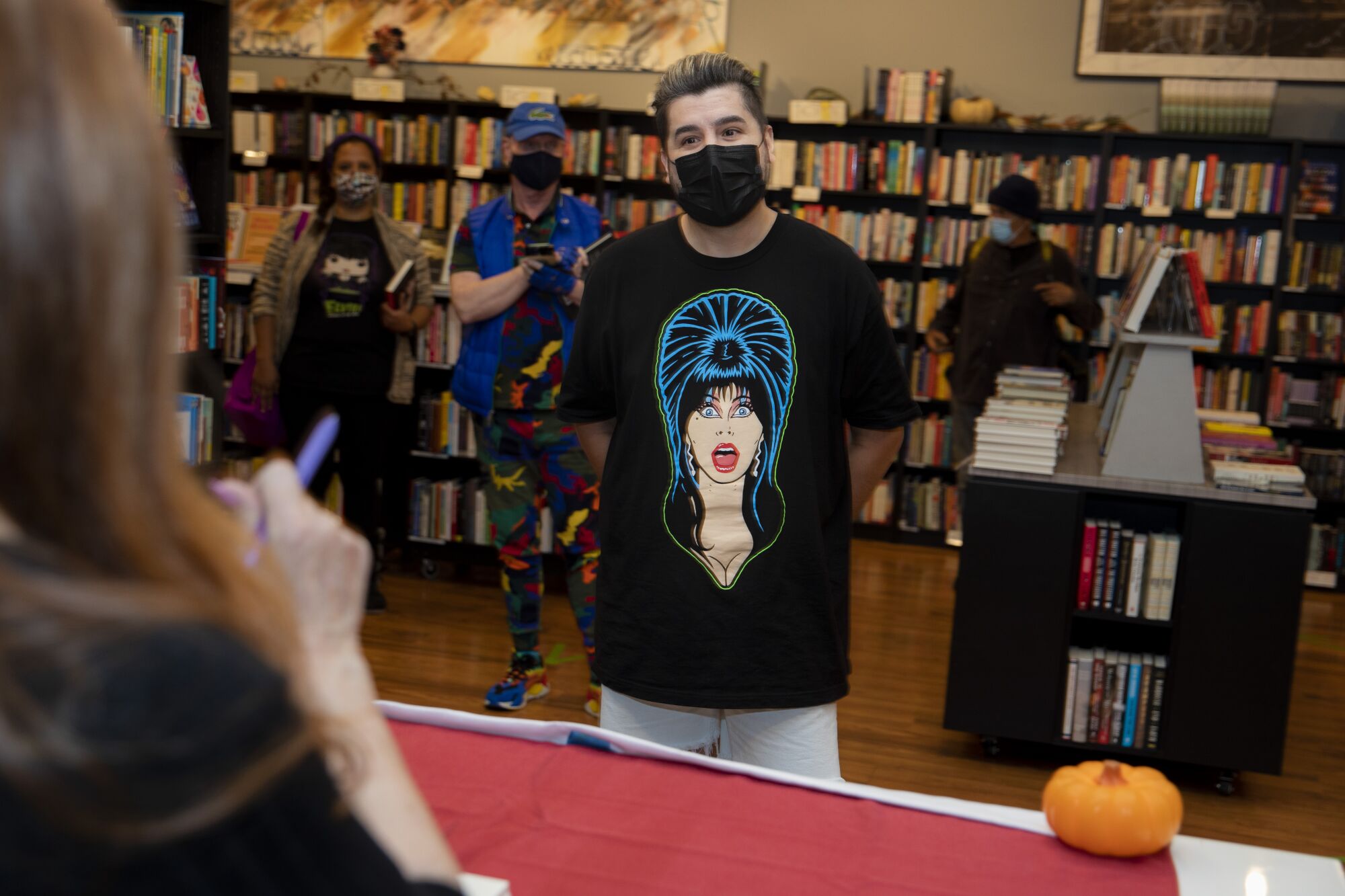 A man wearing a mask and a T-shirt of Elvira stands in line
