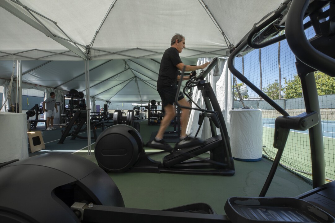 مردی در یک سالن ورزشی در فضای باز زیر یک چادر با یک دستگاه بیضوی در فضای باز تمرین می کند. 