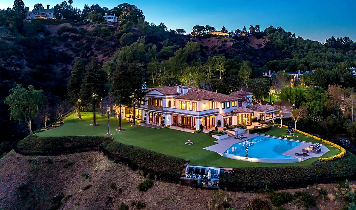 A mansion and pool are surrounded by lawn and trees on a hill.