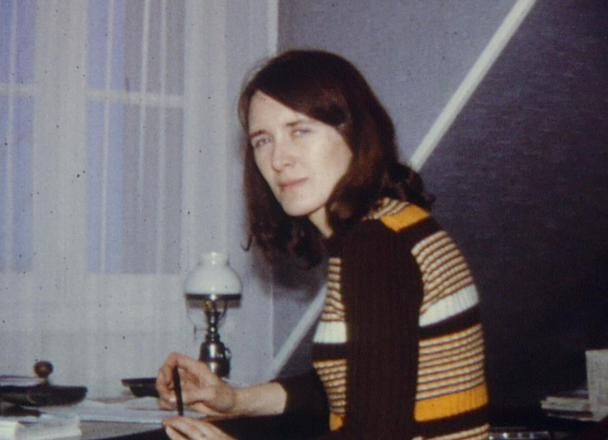 A woman seated at a desk, pen in hand, turns to look at the camera.