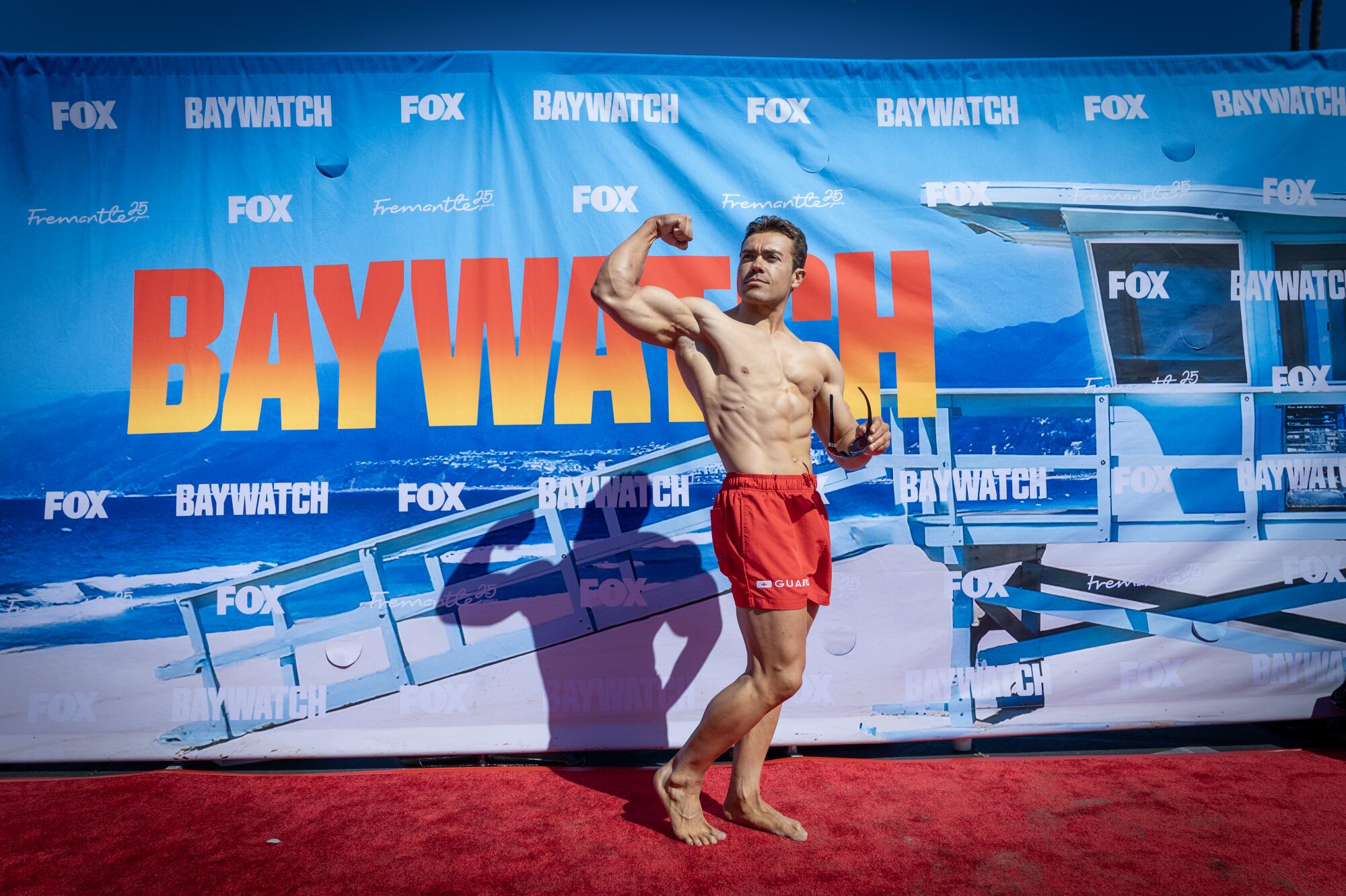 Um homem musculoso flexionando-se em um tapete vermelho com uma faixa que diz Baywatch.