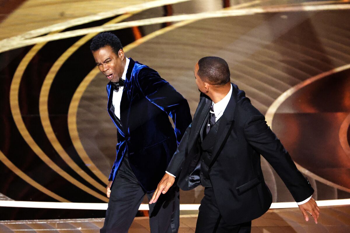 Two men in suits stand onstage. One recoils from the other slapping him.
