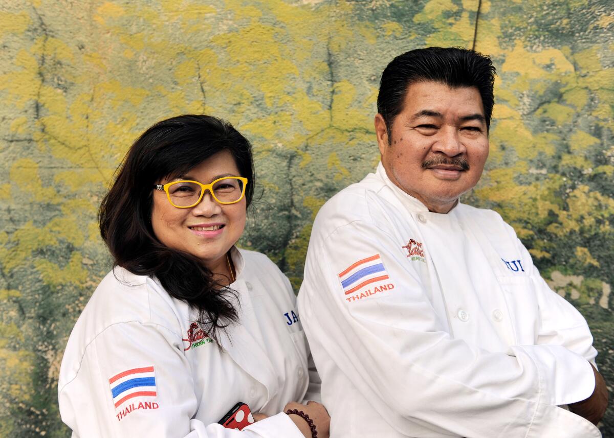 A woman at left and a man, framed waist up, smile and look at the camera, both wearing white chef's coats.