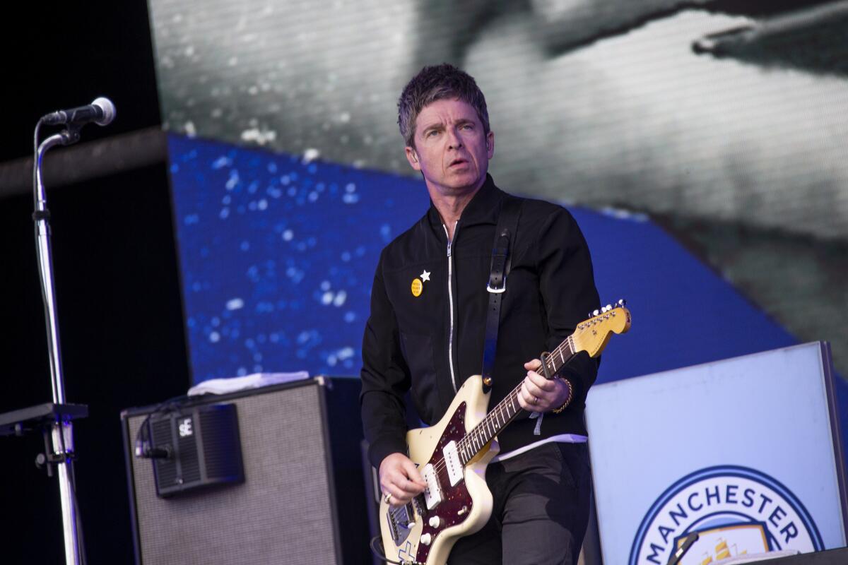 Noel Gallagher, in black jacket and pants, plays a cream-colored guitar onstage.