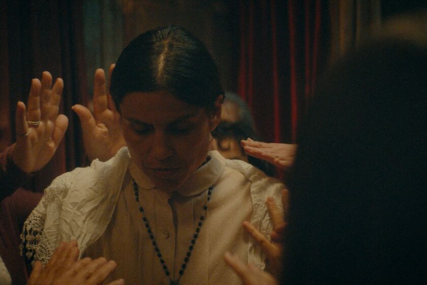A woman in white with her head bowed is surrounded by people with raised hands during a ritual in the movie "Clara Sola."