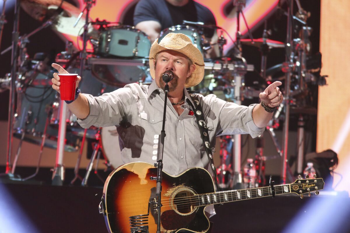 A man in a cowboy hat with a guitar holding a red solo cup while singing into a microphone